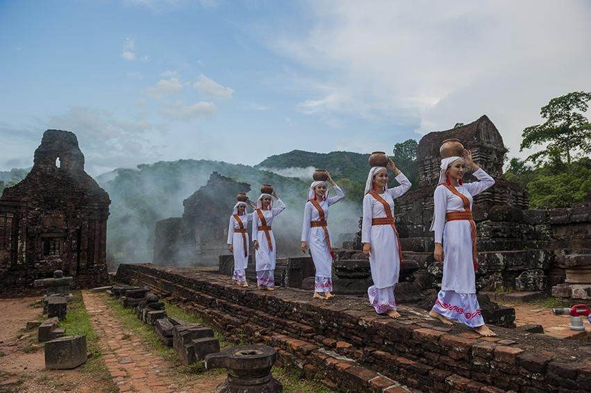 A group of women in white dresses with orange sashes holding pots on their heads Description automatically generated